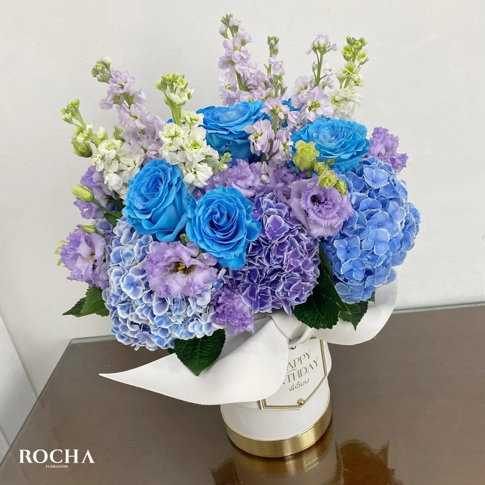 Rocha Floraison hat-box arrangement of blue roses, hydrangeas and lilac stocks with a white satin ribbon